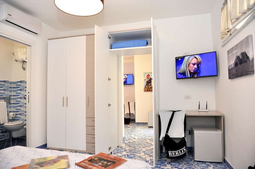 A modern bedroom with white built-in wardrobe, wall-mounted TV, and en-suite bathroom visible through the open doorway.