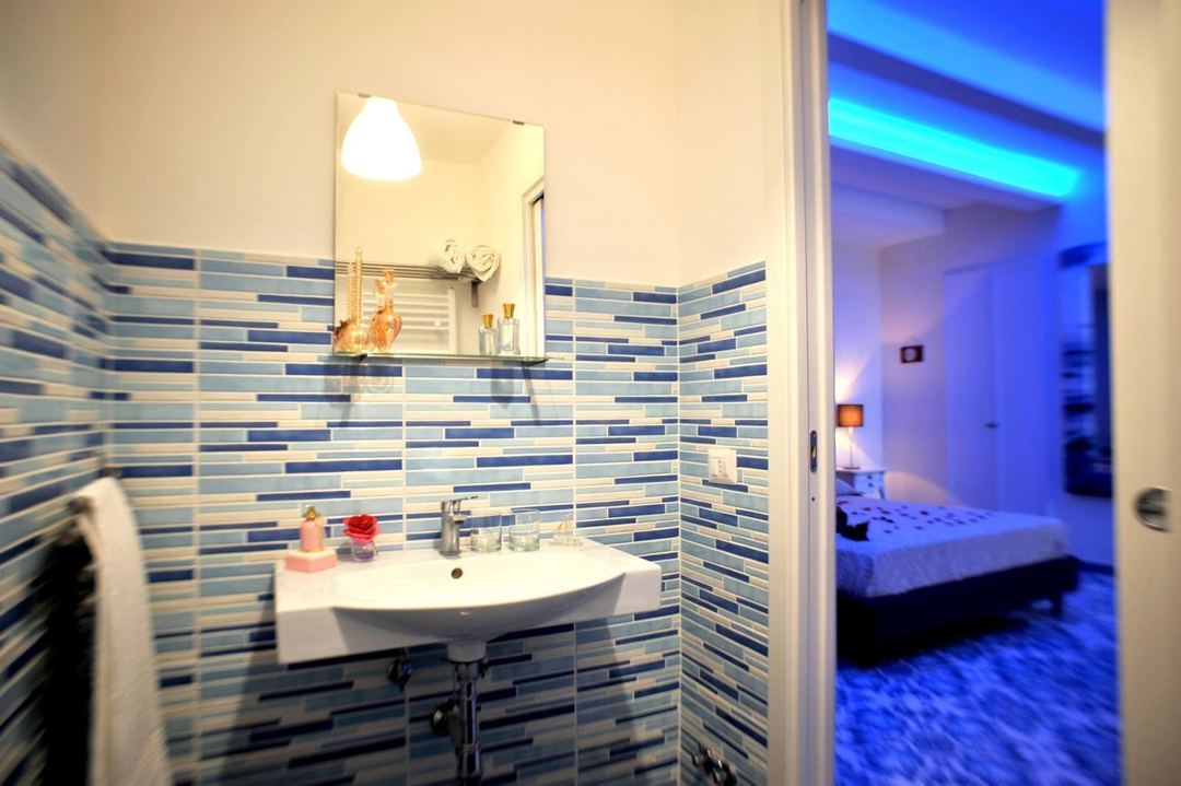 A modern bathroom vanity with blue and white striped tile backsplash, illuminated mirror, and view into an adjacent bedroom.
