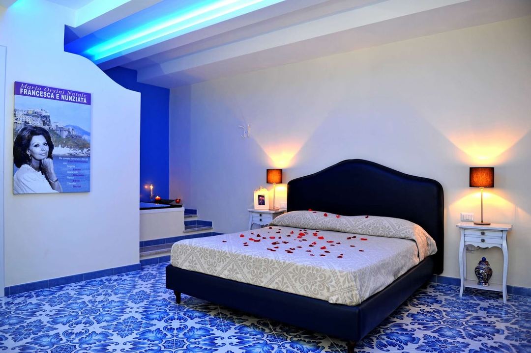 A luxurious bedroom suite featuring a black upholstered bed with decorative rose petals, white nightstands with warm lighting, and blue and white tiled flooring with ornate patterns.
