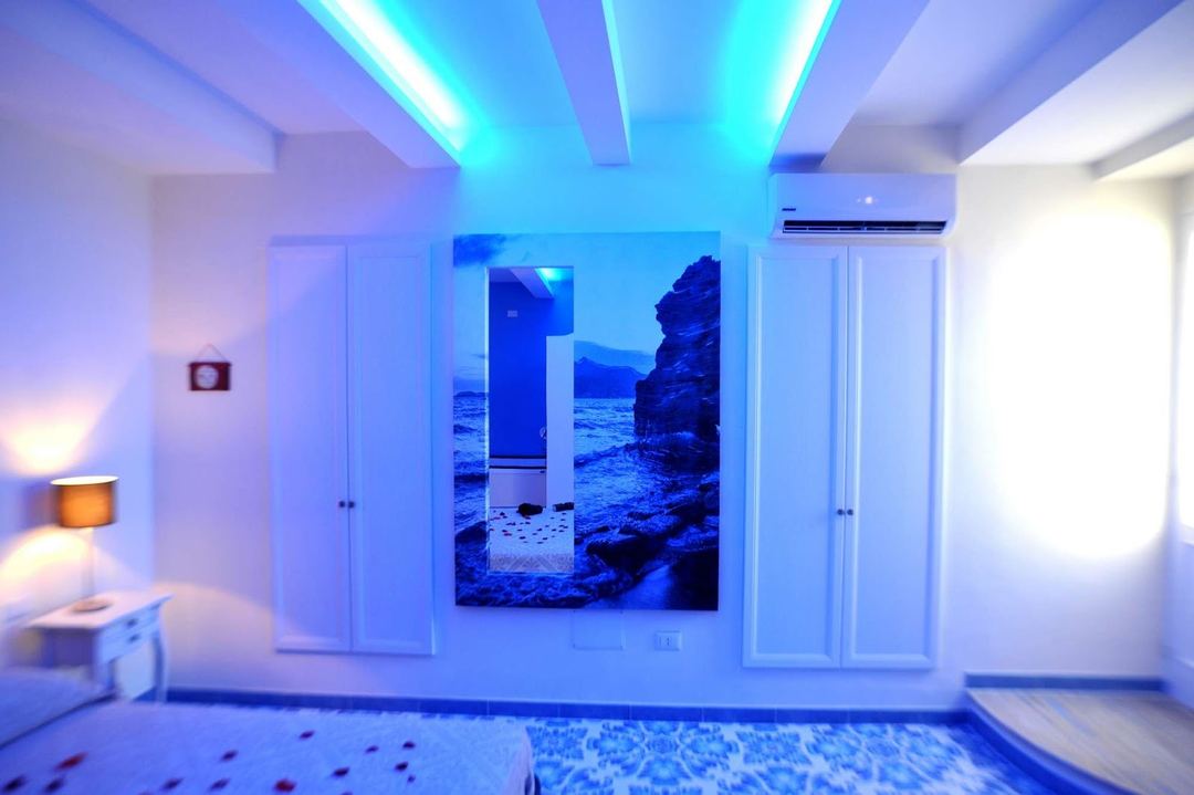 A modern bedroom with blue ambient lighting, white built-in wardrobes, and a coastal landscape artwork on the wall.