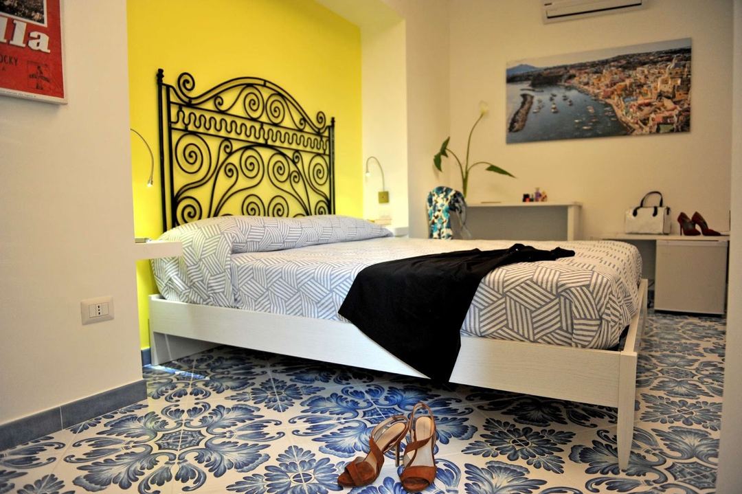A bright bedroom featuring a white bed with geometric patterned bedding, an ornate black iron headboard, and a striking yellow accent wall.