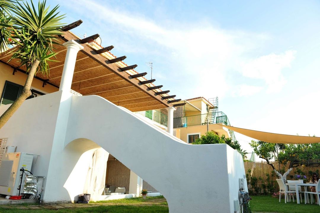 Modern white Mediterranean-style building with wooden pergola, palm tree, and manicured garden grounds.
