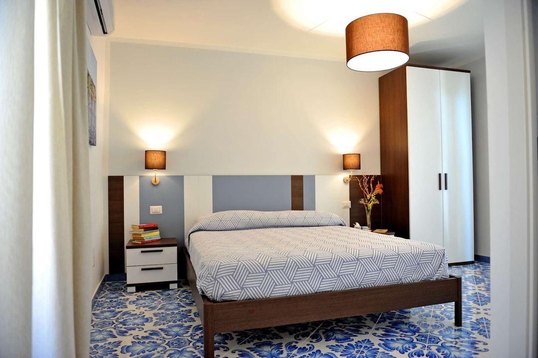 A modern bedroom featuring a wooden bed frame with patterned bedding, slate blue headboard, white nightstands with warm lighting, and a decorative blue patterned floor.
